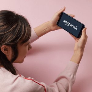 Pink background. Photo of an asian woman laying down holding a smartphone with the Amazon Ads logo displayed.